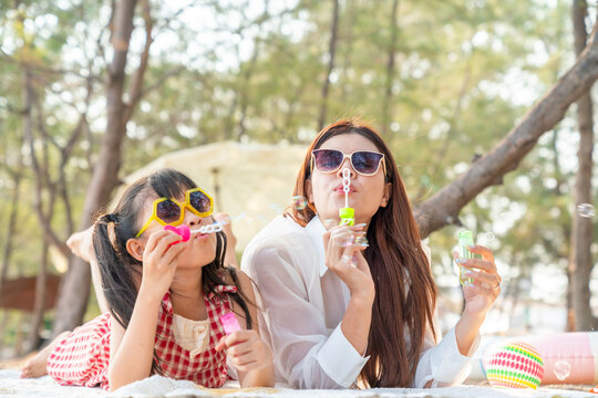 Happy Asian mother and little daughter blowing soap bubbles on beach blanket by the sea, family travel outdoor lifestyle on summer holiday vacation, parent and child bonding spending time together.
