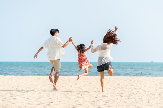 Happy Asian family parents and child playing and jumping together at tropical beach. Father, mother and little daughter enjoy outdoor active lifestyle travel the ocean on summer holiday vacation.
