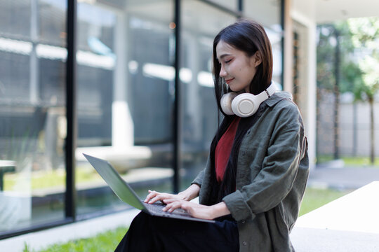 Young asian woman student smiling, concentrating while typing on laptop computer outdoors, enjoying focused remote work and online learning with wireless headphones