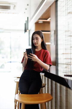 Young asian woman in a red shirt using her smartphone in a modern coffee shop, embodying digital communication, social media use, and connected lifestyle in an urban environment