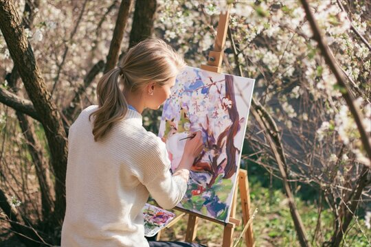 Young female artist paints blossoming trees outdoors