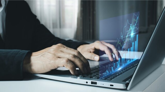 Side view of corporate businessman typing on laptop keyboard with glowing transparent digital financial graph bar projection chart
