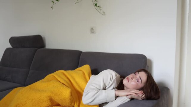 Young woman resting on sofa under warm blanket while experiencing fever weakness and exhaustion during viral infection recovery spending the day isolated at home