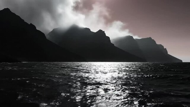 Dramatic seascape of dark mountains with mist and shimmering water under moody skies evokes a sense of wild isolation
