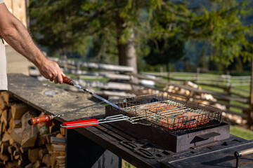Fototapeta premium Man cooking meat on outdoor barbecue. Man uses a metal skewer while grilling meat on a charcoal barbecue in a sunny rustic mountain yard.