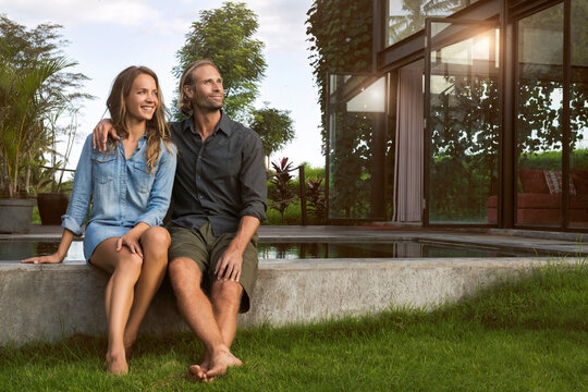 Smiling hugging couple sitting on the edge of a pool in front of their design house