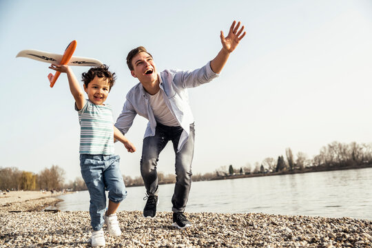 Playful man and boy holding airplane toy while running by lakeshore on sunny day