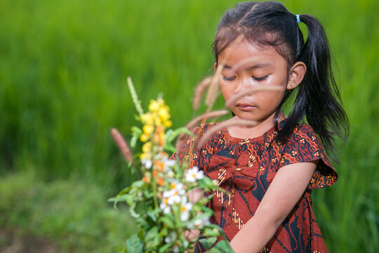 A young Asian girl in rural Indonesia wearing a traditional brown batik dress with parang motifs and a bow accent.