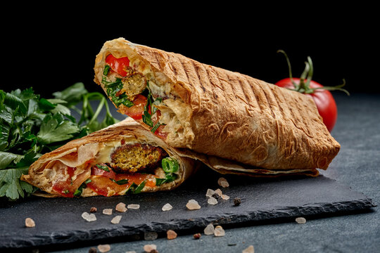 Gourmet vegan shawarma wrap cut in half with plant-based meat and fresh vegetables on black slate board, side view of delicious meatless burrito or doner kebab on dark background
