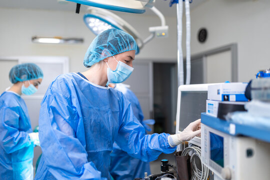 Profile of focused female anaesthesiologist in blue sterile gown, mask, and cap, using gloved hand to adjust medical monitor in operating room, blurred surgical team background. Healthcare concept.