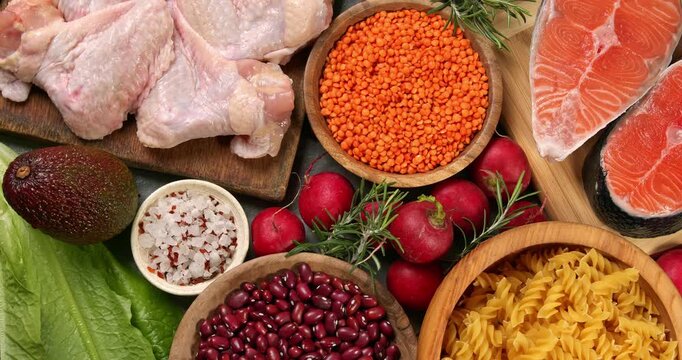 Healthy products on a gray table. Close-up footage of fish, meat, chicken, onion, radish, lettuce, mint, avocado, tomatoes, potatoes, eggs, rice, carrots, beans, lentils, macaroni, corn porridge.