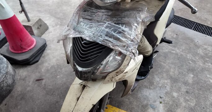 close up of a mud splattered scooter, with its headlight partially covered by makeshift plastic sheeting, waits at a gas station to refuel