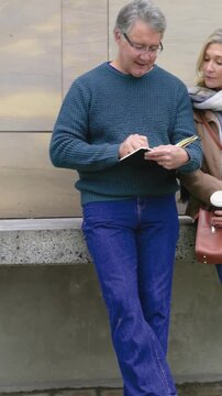 Vertical video: Opening notebook, man in teal writing at plaza, woman holding coffee viewing notes