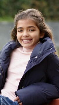 Vertical video: Smiling girl in pink top navy puffer leaning on bench, camera zooming meeting gaze