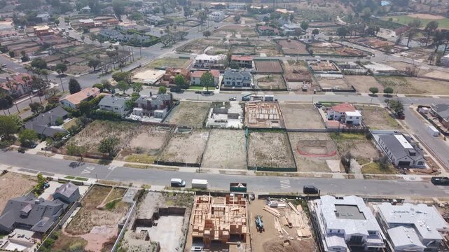 Pacific Palisades wildfire aftermath with rows of empty lots and active construction frames, surviving homes and dry hazy hills visible beyond under pale sky. California, USA. Aerial flyover shot