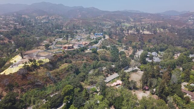 Pacific Palisades burned hillsides with yellow erosion tarps and scattered surviving homes among scorched trees, dry mountains rising beyond under hazy sky. California, USA. Aerial flyover.