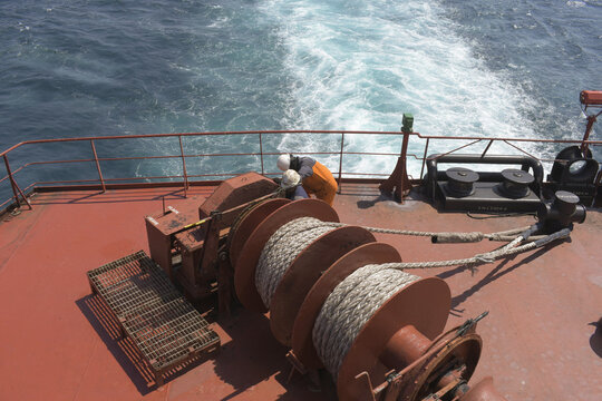 crew members working on mooring winches and ropes at the stern deck of a cargo ship sailing through open ocean in bright daylight