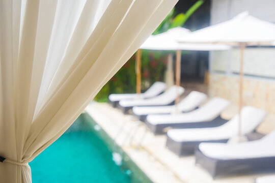 Private cozy resort swimming pool with lounge chairs and umbrellas behind white curtain in focus. Luxury summer vacation background.
