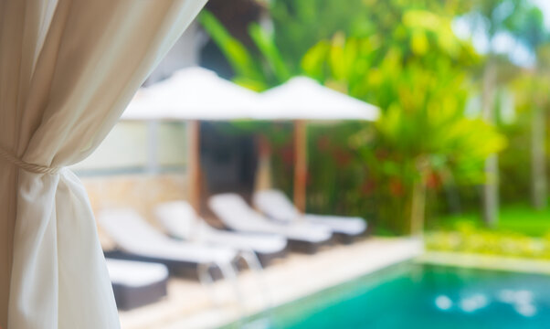 Private resort swimming pool with lounge chairs and umbrellas behind white curtain in focus. Luxury summer vacation background.
