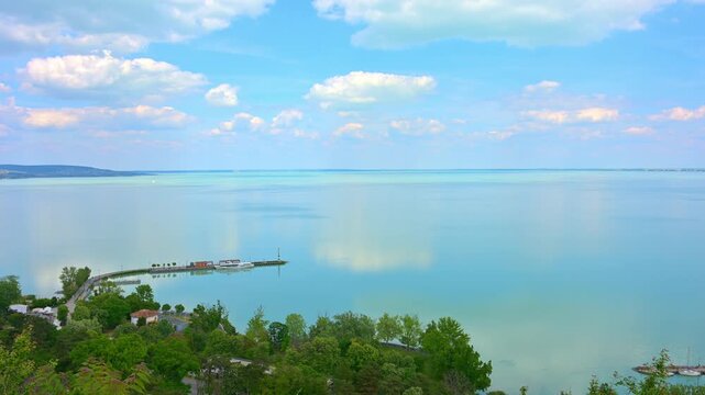 View from Tihany overlooking the calm waters of Lake Balaton in Hungary. Scenic landscape featuring cloud reflections, lush greenery, and a small harbor on a bright spring day.