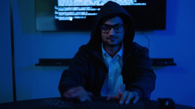 Excited asian young male hacker wearing a hoodie types on a computer keyboard in a dark blue lit room. The talented programmer celebrates digital success after writing code. Cyber security concept.