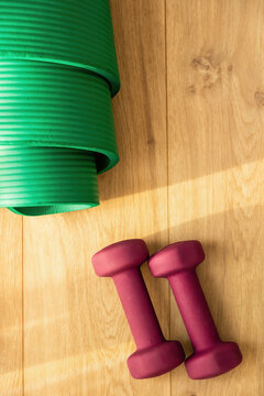 Minimalist flat lay of purple dumbbells and a green fitness mat on a wooden floor in sunlight. Ideal for home workout and wellness content