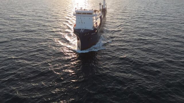 High-angle drone shot showing a cargo ship slicing through the water at sunrise, focusing on its bow and wave formation.