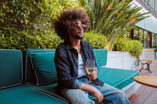 Young man with afro hair enjoying a glass of white wine on a trendy hotel patio.

