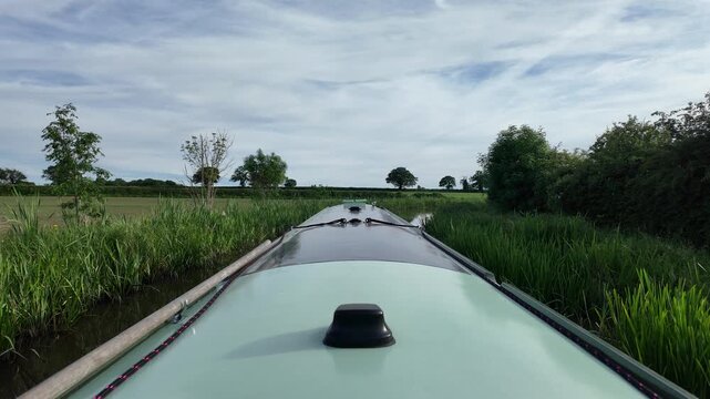 Looking down the centre of a green narrowboat cruising along a canal. The canal has lush greenery to the side, and the water is calm. The scenery is rural, beautiful and calming.