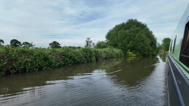 Looking along the side of a green narrowboat cruising along a canal. The canal has lush greenery to the side, and the water is calm. A historic canal bridge is approaching in the distance.