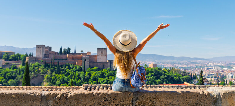 happy woman tourist with arms raised sitting on stone wall looking at Ahambra fortress in Granada, Andalusia in Spain