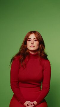 Redhead woman in a red turtleneck dress sits against a green screen backdrop and smiles calmly.