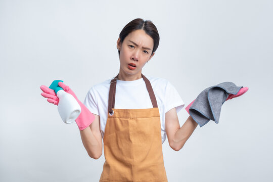 Asian woman in an apron and pink rubber gloves, looking confused and holding cleaning tools, isolated on white background. Concept of housework problems and cleaning challenges.