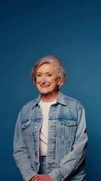 Portrait of a happy senior woman in denim jacket and white t-shirt, standing against a vibrant blue studio backdrop, with a warm, welcoming smile.