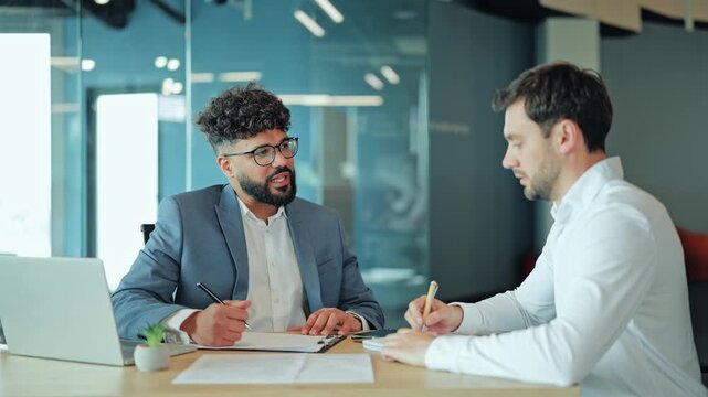 Two male partners in formal shirts and suits sitting at a wooden desk in a contemporary glass office. Caucasian and Arab men seriously signing important business documents and contract with pens.