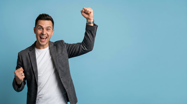 Ecstatic young man shouting with joy and raising his fists in celebration of success isolated against a plain blue background.
