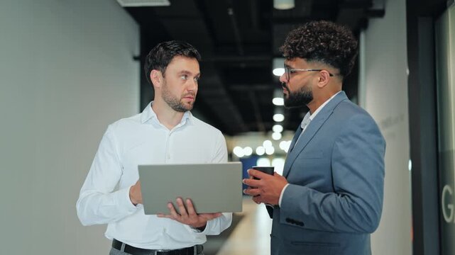 Businessmen standing in modern corridor and analyzing presentation data on laptop. Multiethnic team members working together on corporate project during coffee break.