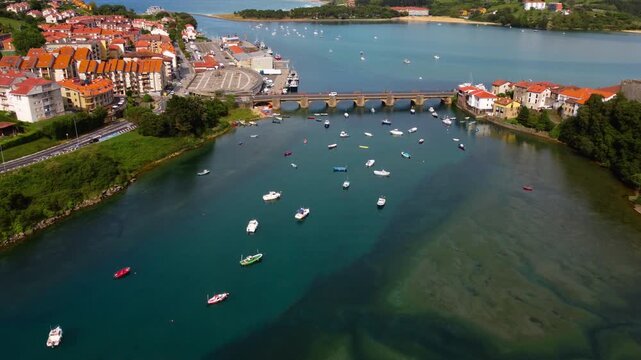 Top down aerial view of Barquera Bridge in San Vicente de la Barquera, showing colorful fishing boats moored in the turquoise estuary water in Cantabria, Northern Spain. High quality 4k footage