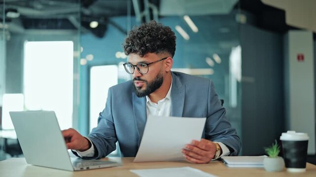 Focused Middle Eastern businessman in glasses holding paper document and verifying information on laptop screen. Serious corporate executive analyzing reports at modern office workspace.