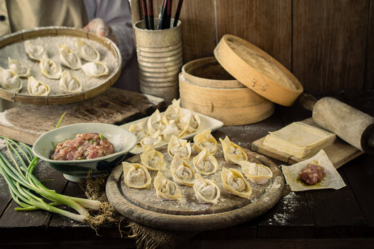 An artisan chef preparing traditional Chinese wontons and dumplings with fresh minced meat, green onions, and dough on a rustic wooden table. Authentic Asian culinary cooking process. 