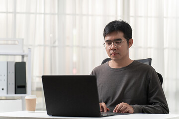 Fototapeta premium Young male professional working on laptop at office desk with beige curtain background