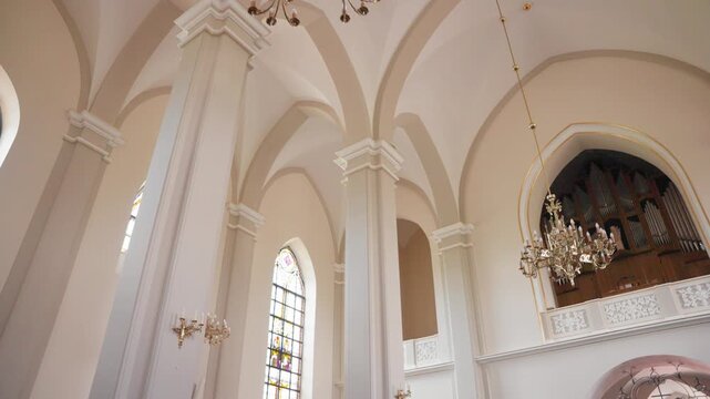 Wide angle glide through a grand church interior with vaulted ceilings, intricate chandeliers, stained glass windows, and a classic pipe organ. Bright, soft natural daylight fills the serene sacred sp