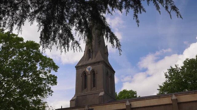 The clock tower of St Leonard's Church rises against a partly cloudy sky, framed by lush green tree branches. Ideal for historical documentaries or architectural features.