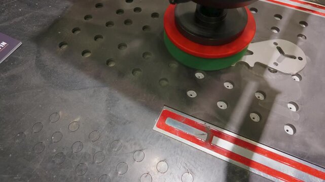 Deburring and linishing machine smooths sharp edges on a metal plate on a perforated workbench, showing abrasive head rotation and surface finishing in a workshop.
