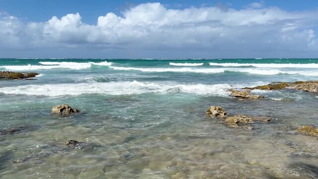 Wide shot of a rugged rocky coastline with turquoise Caribbean waves breaking against the rocks under a clear blue sky with clouds in Puerto Rico. Shot on iPhone 17 Pro.