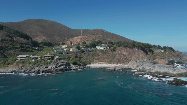 Advancing flight over turquoise cove with rocky shores revealing exclusive clifftop villas nestled into dry scrubland hillside under clear blue sky. Punta Pite, Papudo, Chile