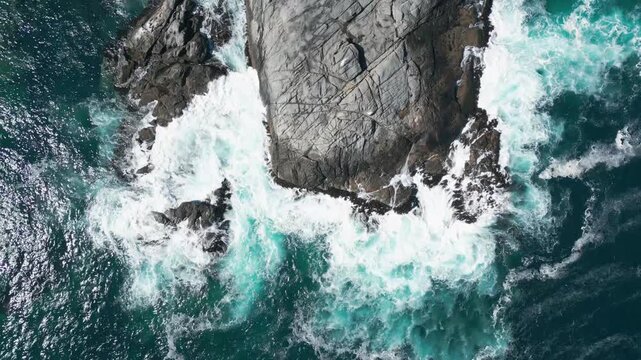 Bird's eye view of large flat rock formation surrounded by turquoise foaming waves and white sea spray against deep blue ocean
