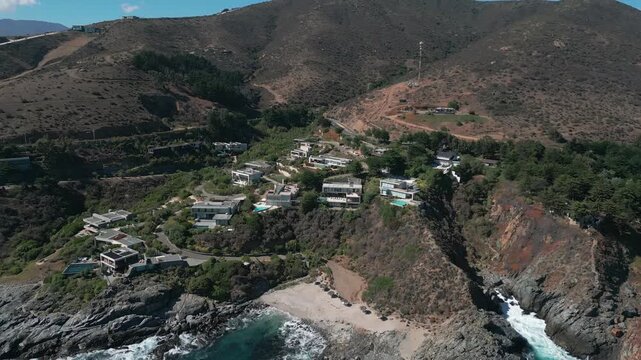 Retreating reveal over exclusive clifftop villas with pools integrated into rugged scrubland hillside above secluded rocky cove and crashing waves. Punta Pite, Papudo, Chile