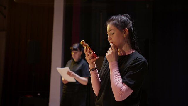 Young performers rehearse lines on stage in a dimly lit theater, reading from phone and paper scripts during an intimate evening acting practice session