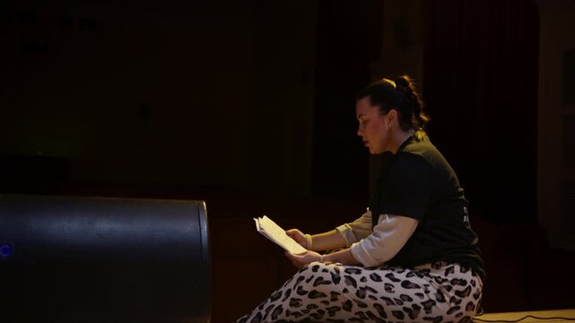 Actor rehearsing alone on dimly lit theater stage with script pages in hand, illuminated by warm spotlight while preparing for an emotional solo performance